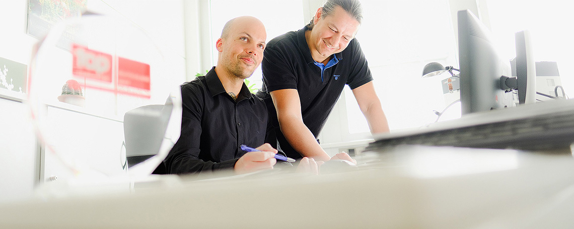 Bild 1: Zwei Kollegen lächelnd bei der gemeinsamen Büroarbeit am Schreibtisch