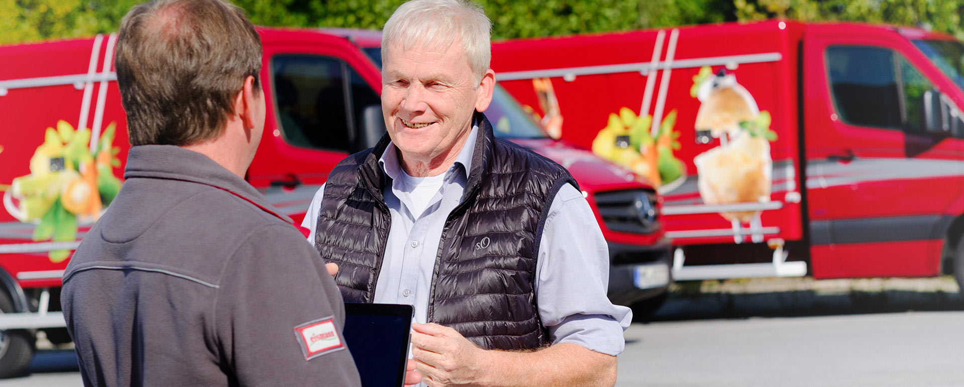 Bild 3: Zwei Männer im Gespräch auf einem Parkplatz vor roten Lieferwagen mit Lebensmittelmotiven