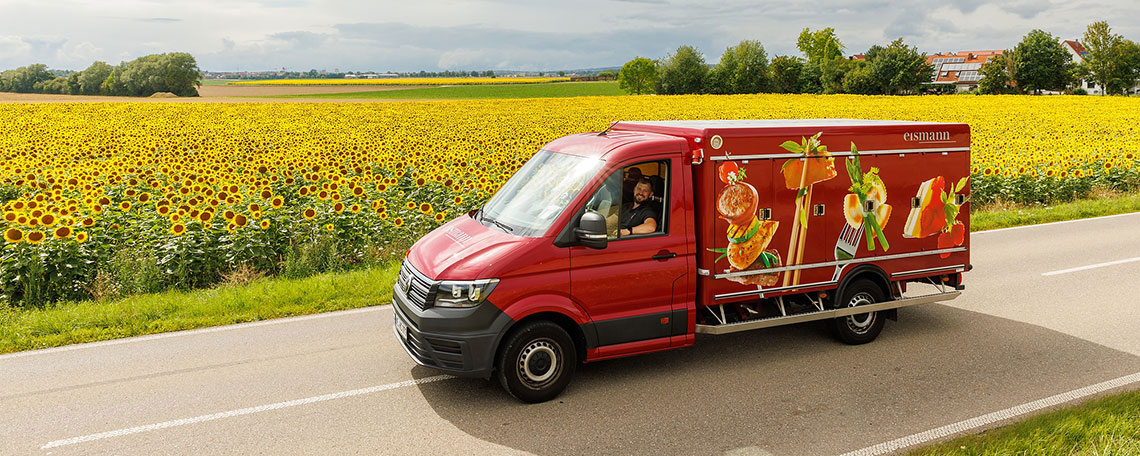 Bild 2: Roter Lieferwagen mit Lebensmitteln-Aufdruck fährt an einem Feld voller Sonnenblumen entlang