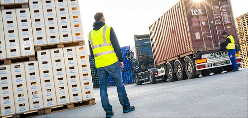 Arbeiter in einer gelben Warnweste steht in einem Lagerbereich neben gestapelten Kisten und einem Lastwagen