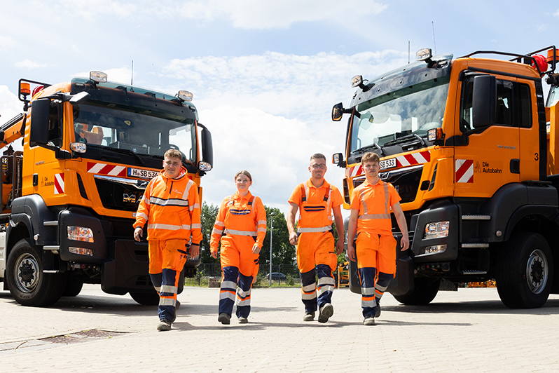Vier junge Autobahnmitarbeiter in orangefarbener Arbeitskleidung stehen vor zwei großen orangefarbenen Lastwagen