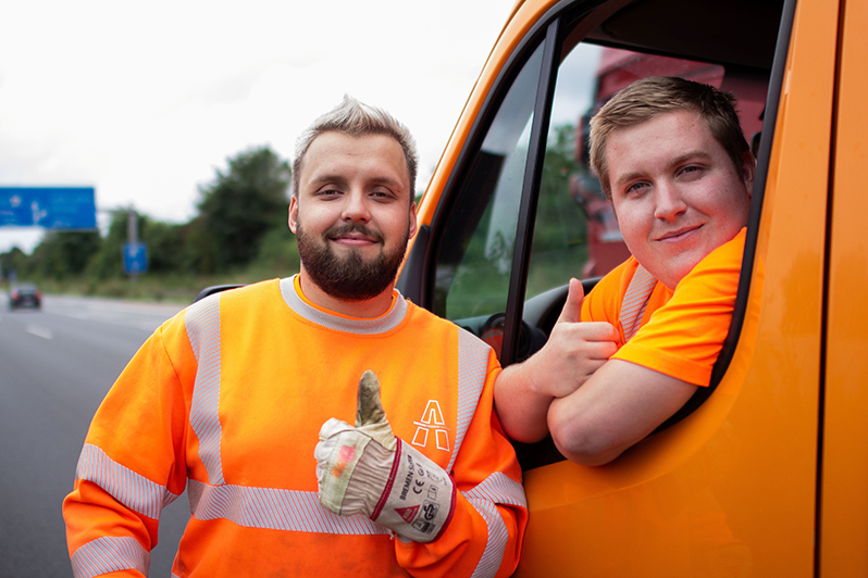 Zwei junge Bauarbeiter in orangefarbener Warnkleidung neben einem orangefarbenen Transporter an einer Autobahn