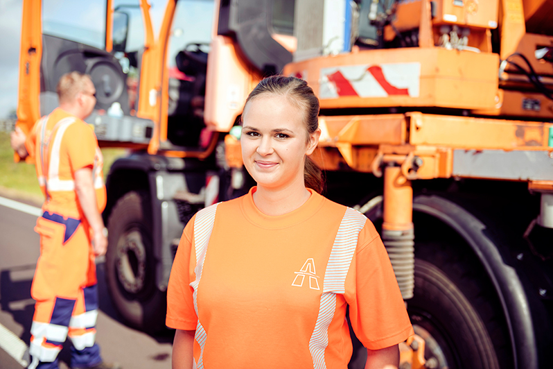 Junge Frau in orangefarbenem Arbeits-T-Shirt mit Autobahn-Logo steht lächelnd vor einem großen LKW