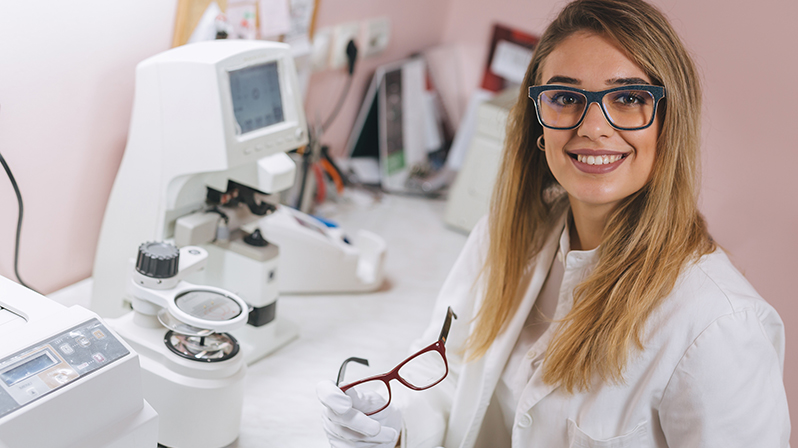 Frau im weißen Kittel hält Brille in Labor mit optischen Geräten