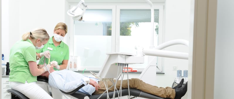 Zwei Zahnärztinnen in grüner Kleidung behandeln einen Patienten im Behandlungsstuhl
