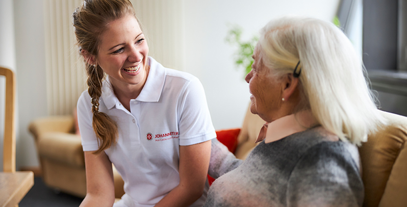 Eine junge Johanniter-Mitarbeiterin lacht herzlich im Gespräch mit einer älteren Dame in deren Wohnzimmer