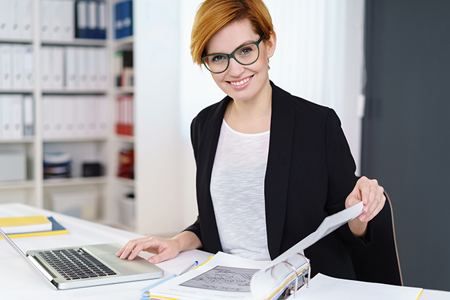 Frau mit Brille am Schreibtisch mit Laptop und Aktenordner
