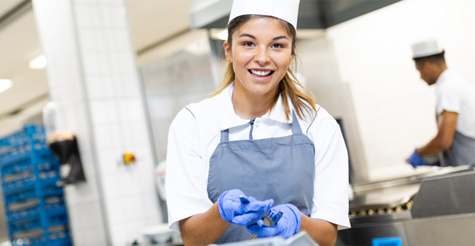 Lächelnde Frau in weißem Shirt, grauer Schürze und blauen Handschuhen in einer Großküche