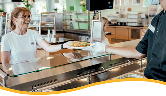 Ein Kellner bedient eine lächelnde Frau im weißen Hemd an der Theke und reicht ihr Essen