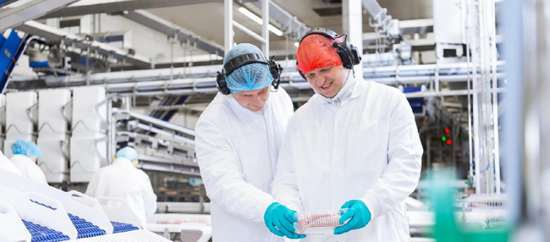 Zwei Mitarbeiter in Schutzkleidung überprüfen ein verpacktes Lebensmittelprodukt in einer modernen Produktionshalle.