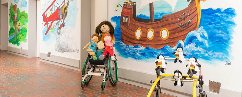 Kindergarten-Flur mit farbenfrohen Wandgemälden. Rollstuhl und Gehhilfe, beide mit Stoffpuppen