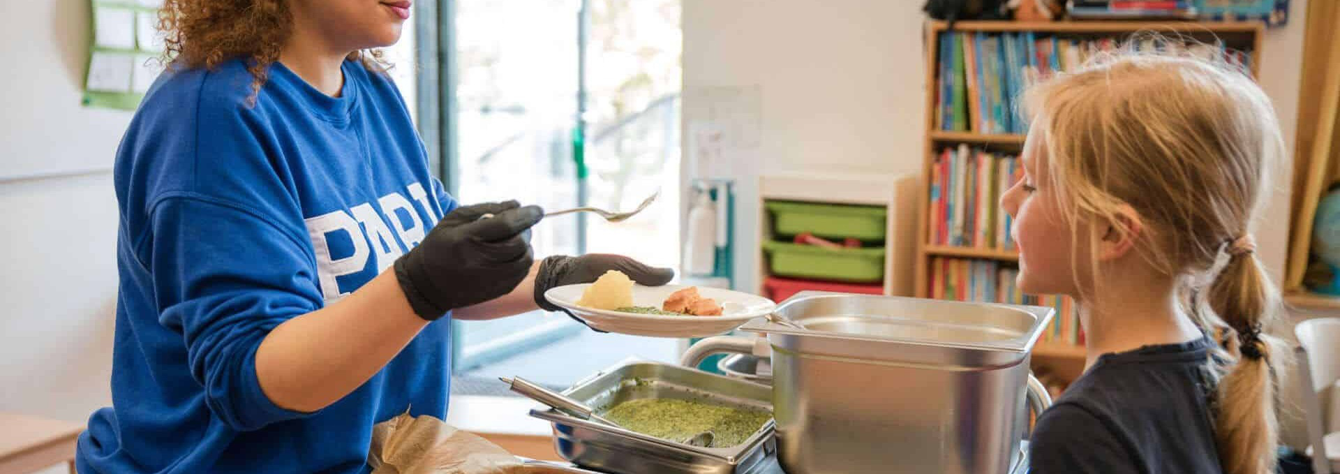 Küchenkraft serviert einem Kind warmes Essen auf einen Teller