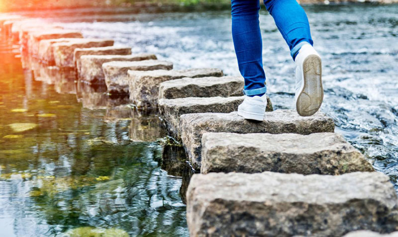 Eine Person in Jeans und weißen Sneakern überquert einen Fluss auf Trittsteinen