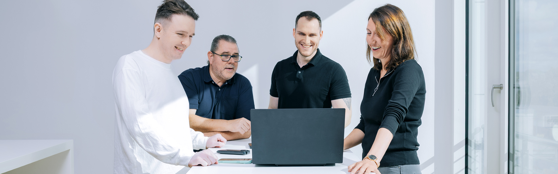Ein Team aus vier Kollegen arbeitet gemeinsam an einem Laptop und lacht in einem hellen Büro