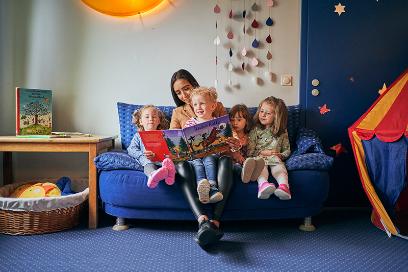 Eine Frau sitzt mit vier Kindern auf einem blauen Sofa und liest ihnen aus einem Bilderbuch vor