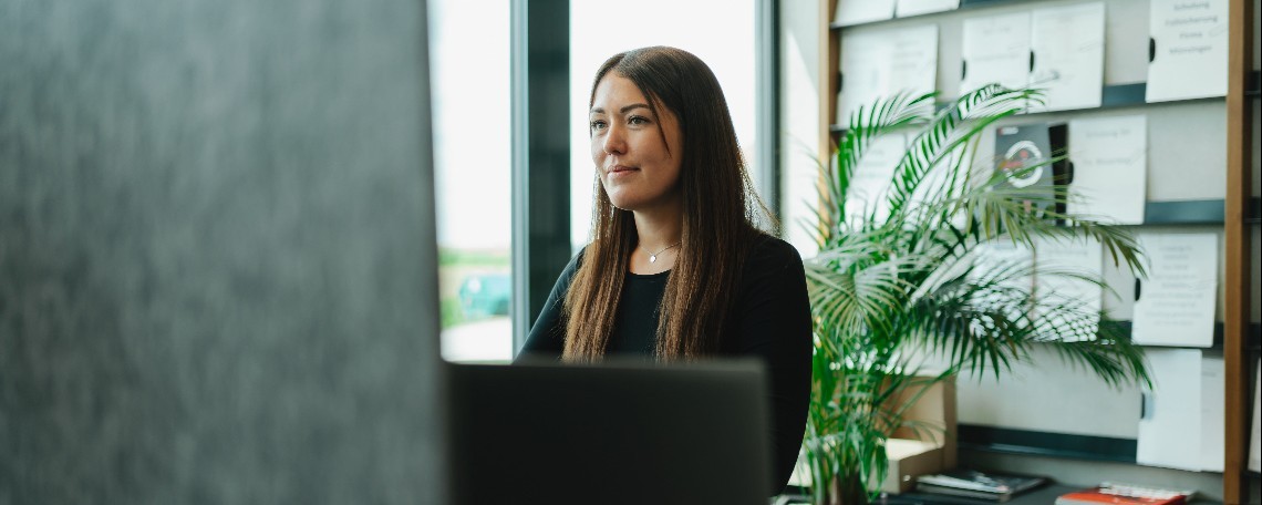 Junge Frau mit langen braunen Haaren arbeitet konzentriert am PC im hellen Büro