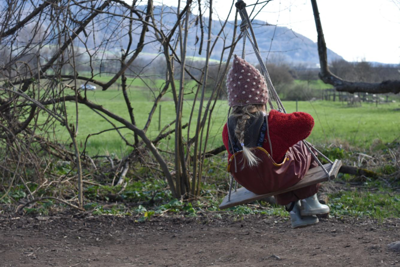 Kind mit Mütze und Zopf schaukelt auf einer Holzschaukel