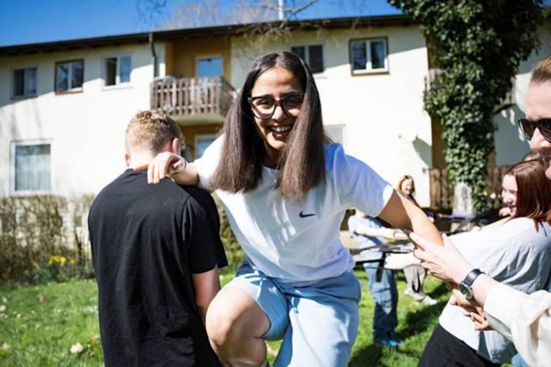 Lachende junge Frau mit Brille wird im Freien von anderen Personen gestützt