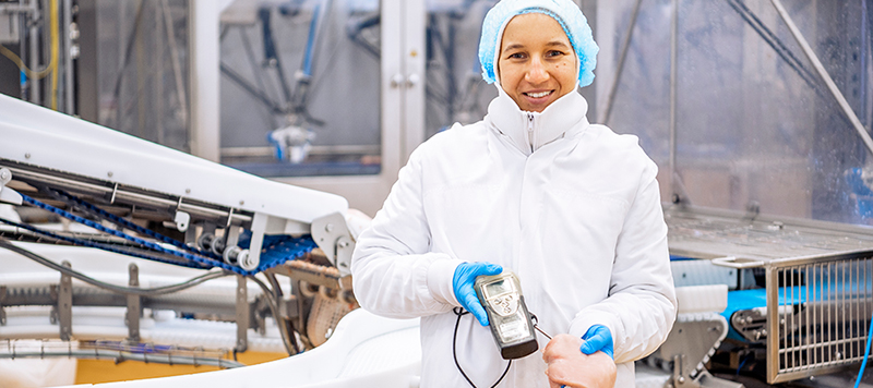 Mitarbeiterin in weißer Schutzkleidung hält ein Messgerät in einer Lebensmittel-Produktionshalle