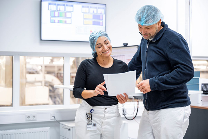 Zwei Mitarbeiter in Schutzkleidung besprechen lächelnd Dokumente in einer modernen Produktionsleitstelle