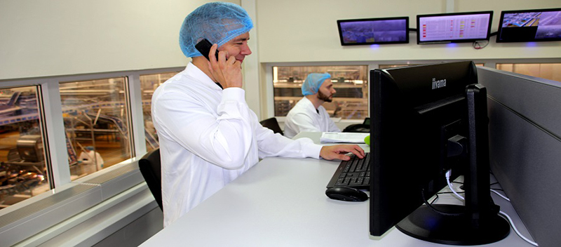 Mitarbeiter telefoniert am PC-Arbeitsplatz in einem Kontrollraum mit Blick in die Produktionshalle