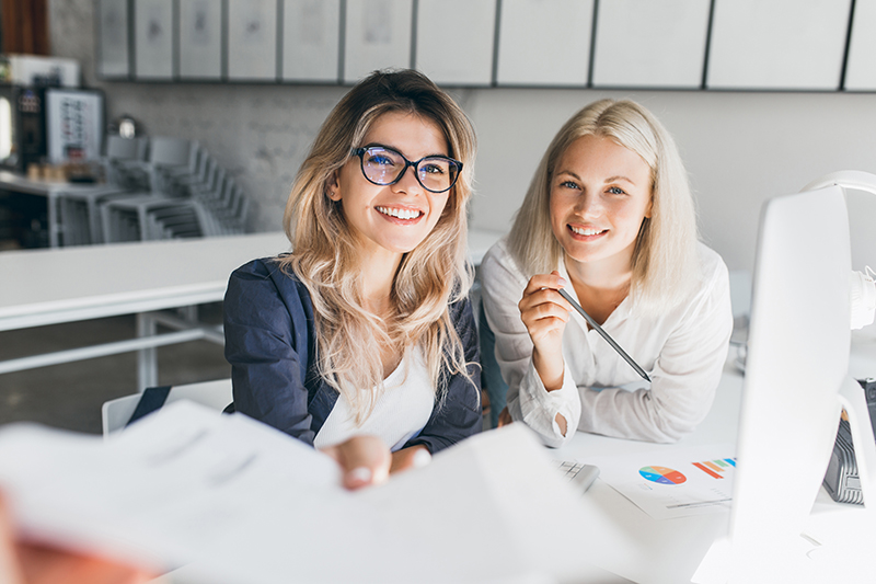 Zwei Kolleginnen im hellen Büro lächeln am Schreibtisch