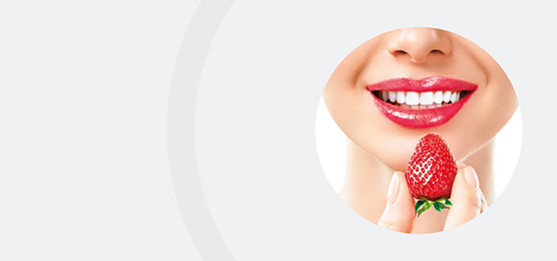 Nahaufnahme eines lächelnden Mundes mit weißen Zähnen und roten Lippen, vor den eine Hand eine frische Erdbeere hält