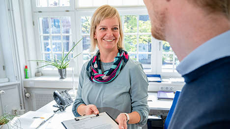 Frau am Schreibtisch reicht einer Person ein Klemmbrett