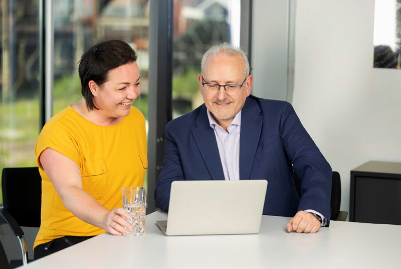 Zwei Kollegen im Büro blicken lächelnd gemeinsam auf einen Laptop